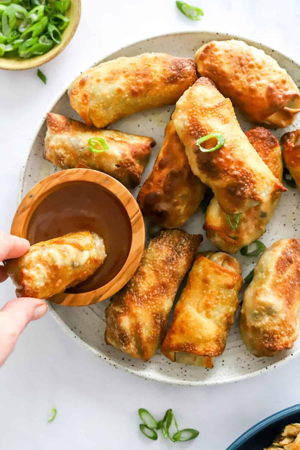 Hand dipping a crispy egg roll into a brown dipping sauce with more egg rolls on the plate around it. 