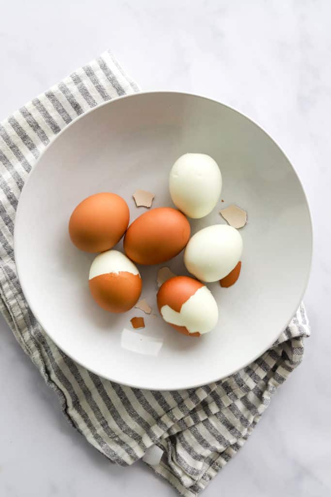 Round white bowl with some whole brown eggs with some peeled in it on top of a striped towel 