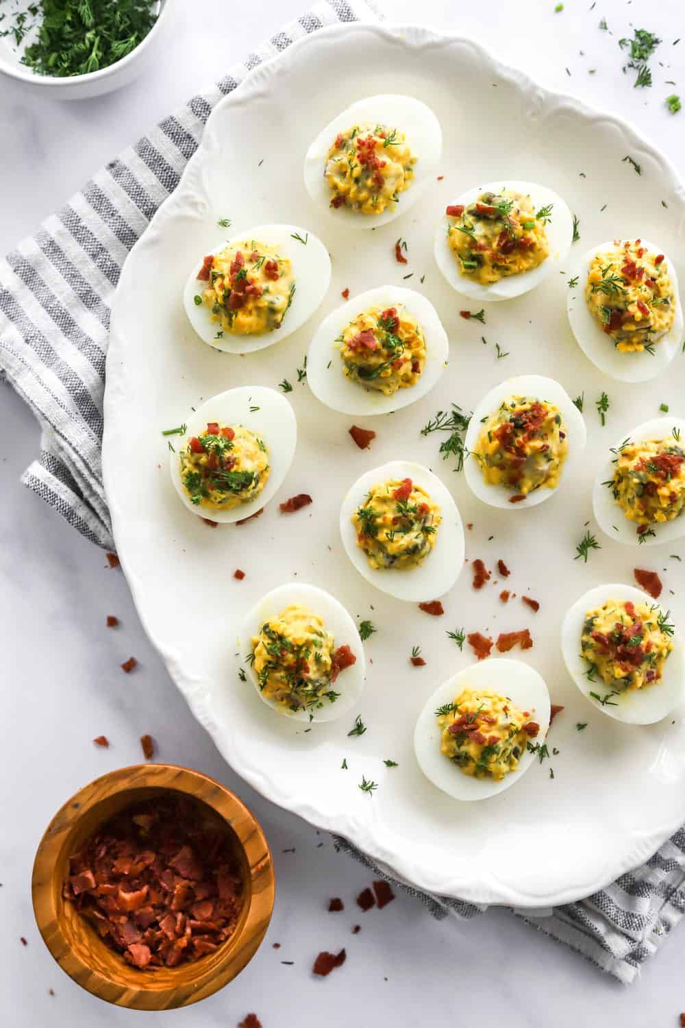Deviled eggs topped with chopped bacon and dill on an oval platter with more bacon in a bowl in front of it and chopped dill behind it on a stripped towel. 