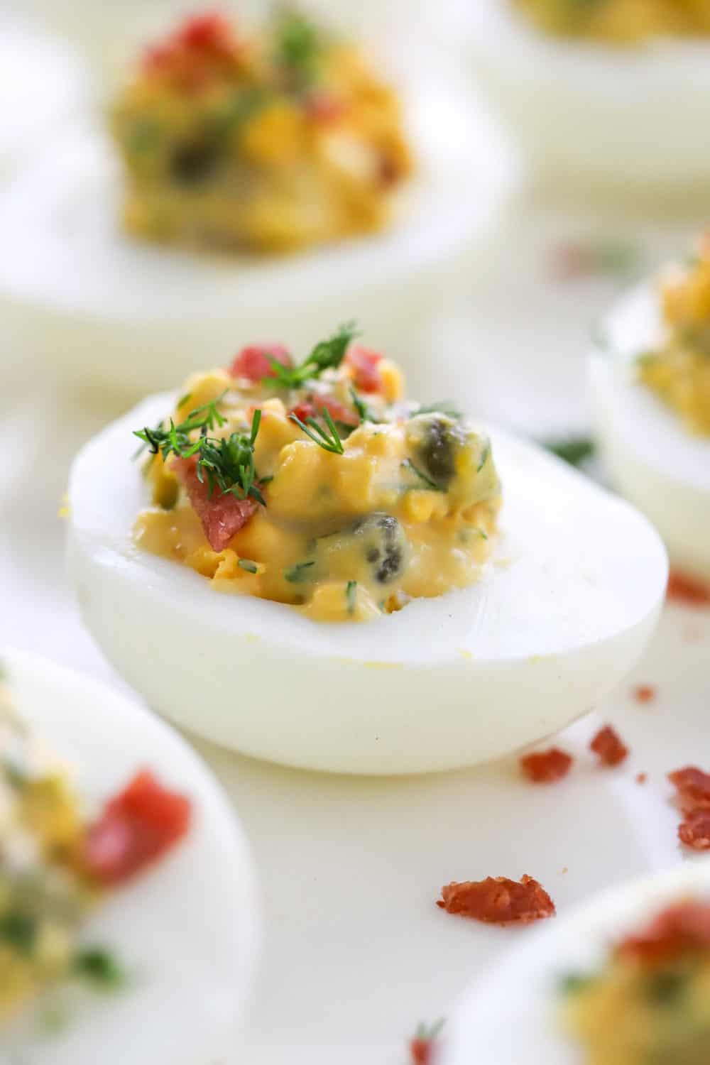 Close up of half of a boiled egg filled with a creamy yellow filling topped with herbs and bacon bits with more eggs around it. 