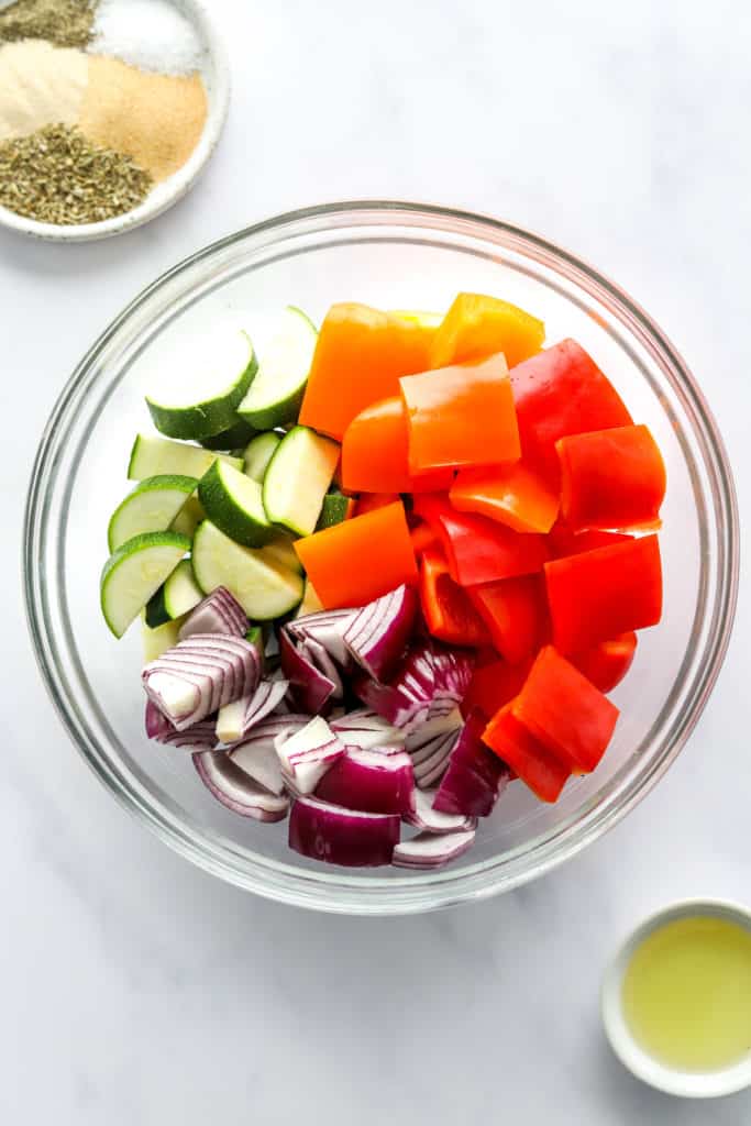 Round glass bowl filled with red and orange bell pepper chopped, plus chopped zucchini and onion with a bowl of oil in front of it. 