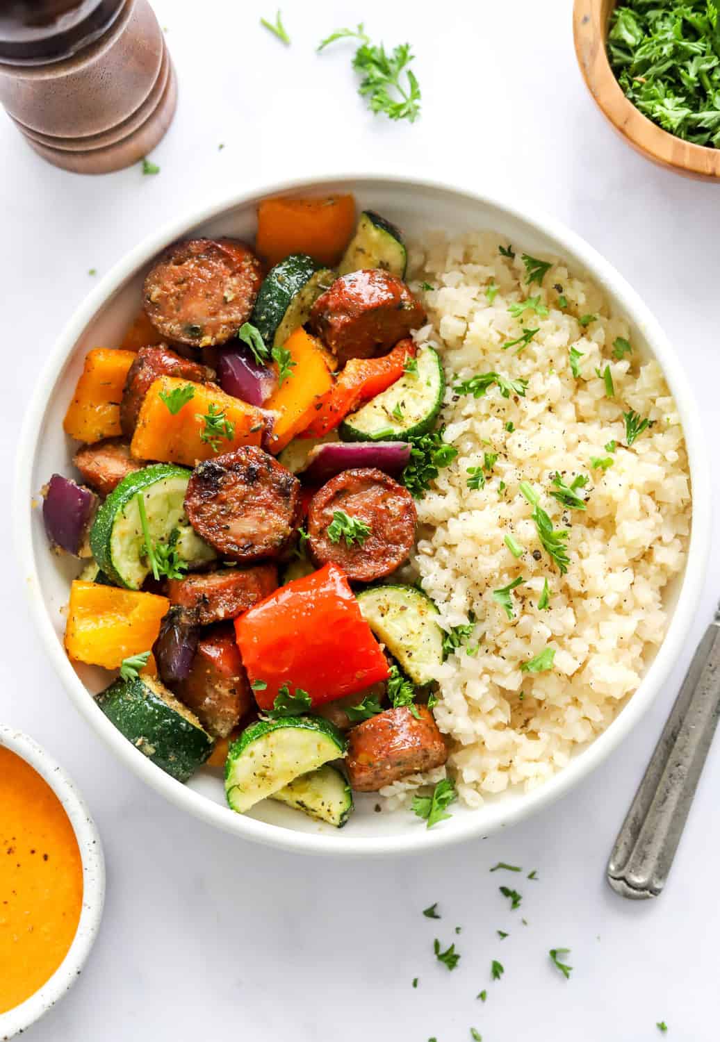 White bowl with crispy sausage pieces and rose peppers and zucchini next to a pile of  riced cauliflower  with a bowl of orange sauce in front of it and a bowl of parsley behind it. 