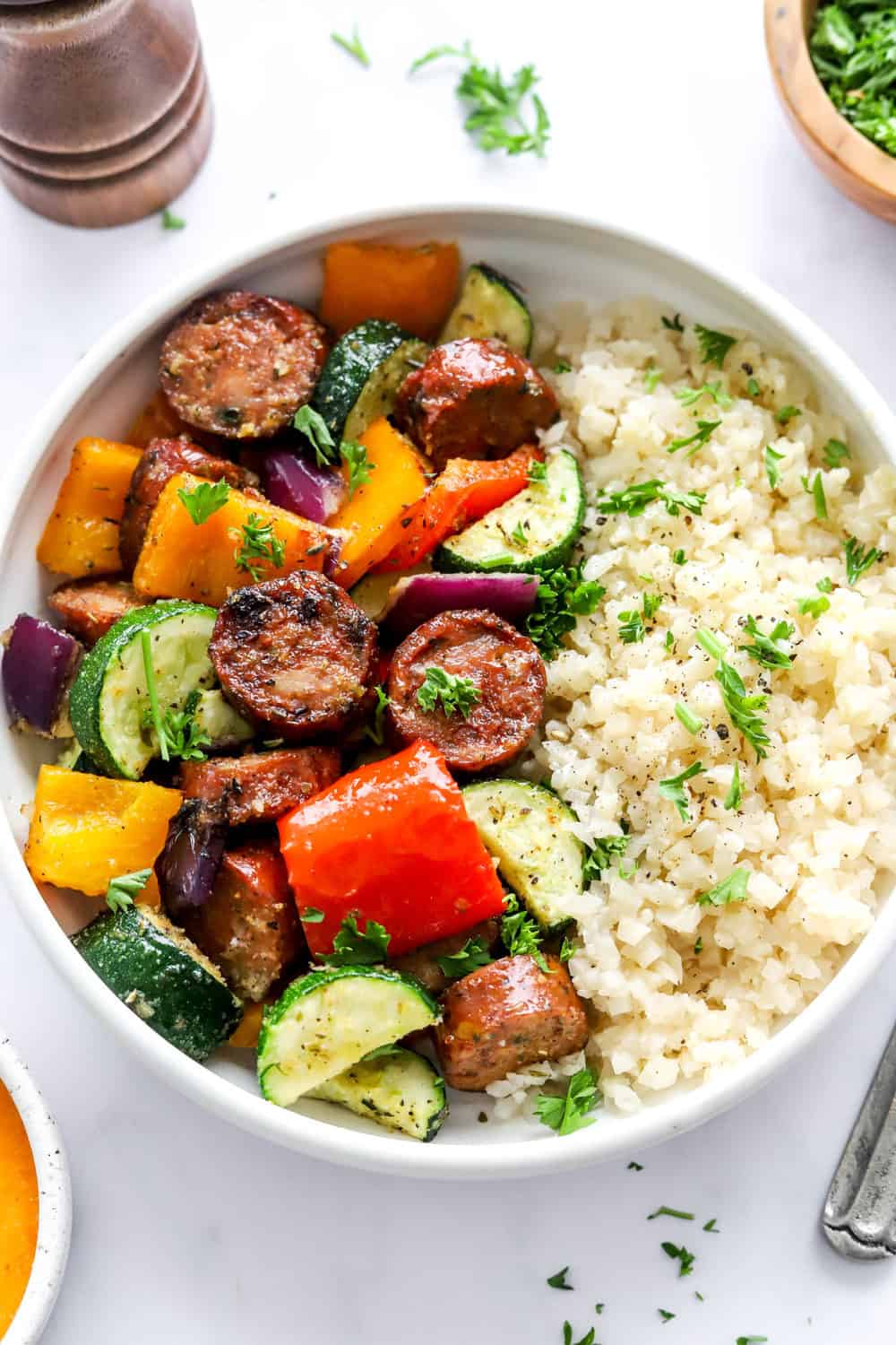 Air Fryer chicken sausage rounds in a bowl with roasted veggies and cauliflower rice with a pepper mill and bowl of chopped herbs behind it. 