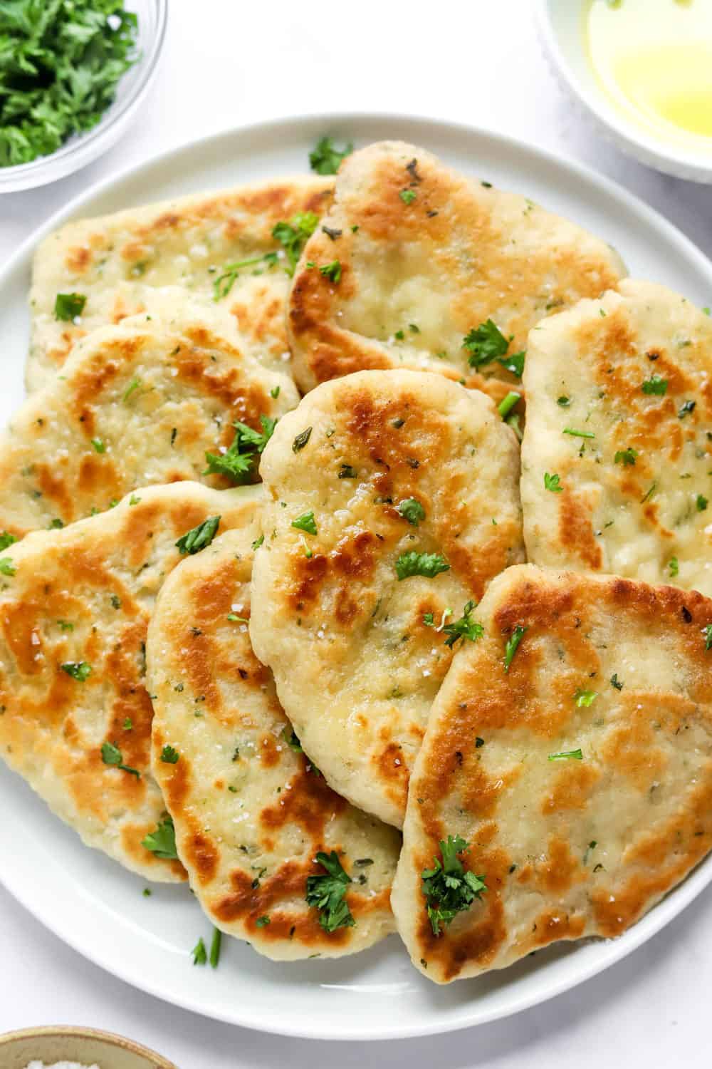 Pile of gluten free naan bread on a white plate topped with some oil and chopped parsley. 