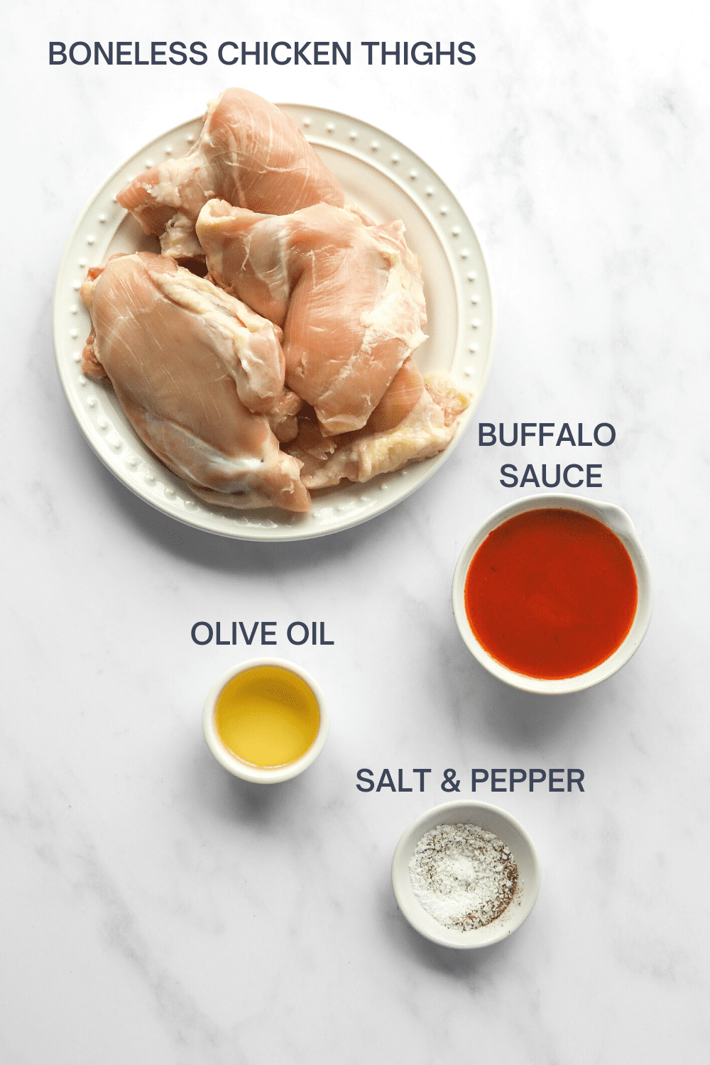 Raw chicken thighs on a round, white plate with a bowl of buffalo sauce, bowl of olive oil and a small bowl of salt in front of it. 