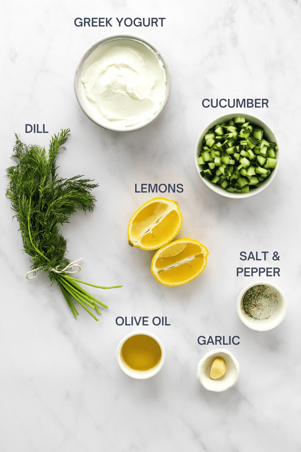 Bowl of plain greek Yogurt with a bunch of dill, bowl of diced cucumber, sliced lemon, small bowl of olive oil, garlic clove, salt and pepper in front of it. 