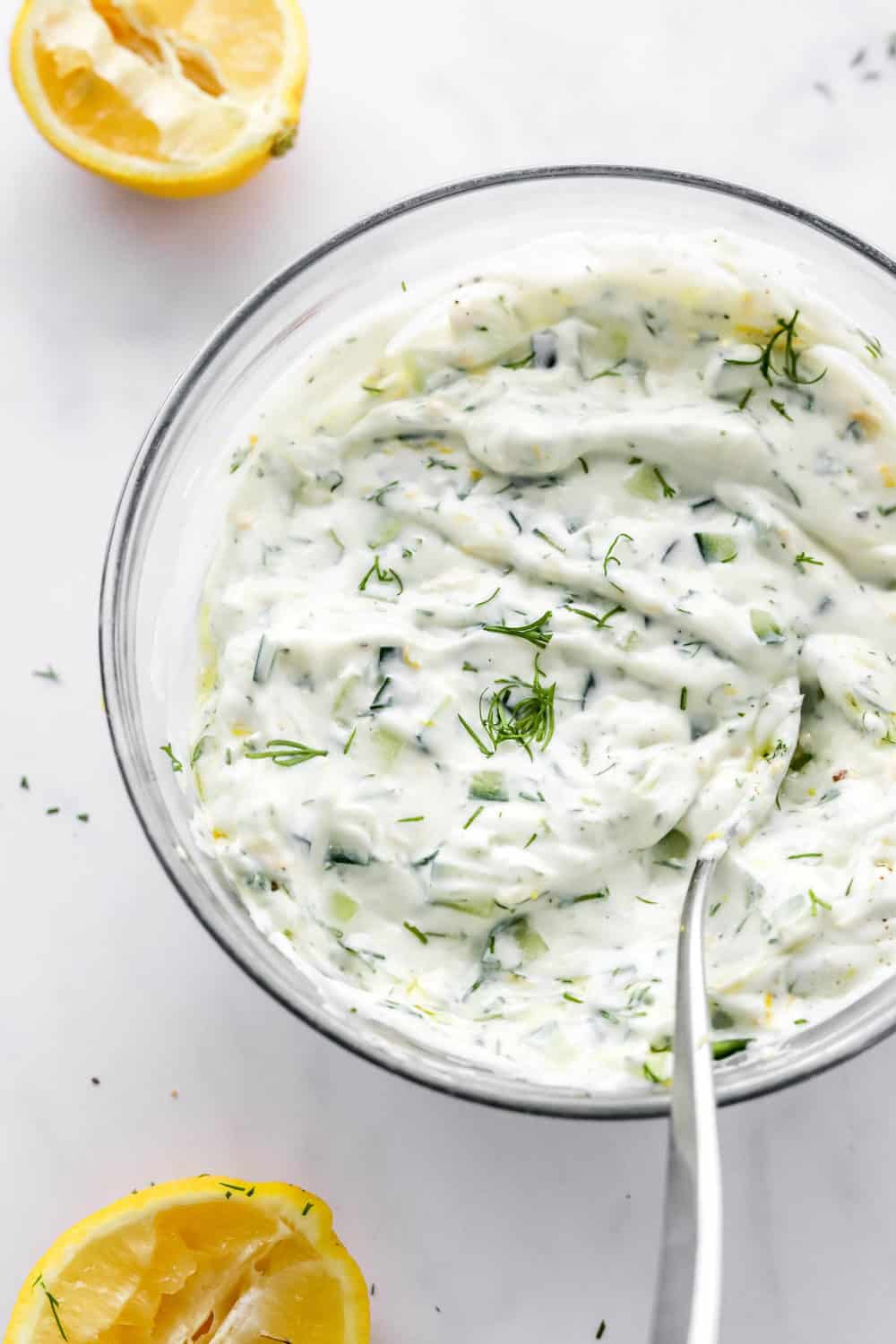 Creamy yogurt sauce with fresh herbs and lemon in a glass mixing bowl with a spoon in it with a sliced lemon in front of it. 