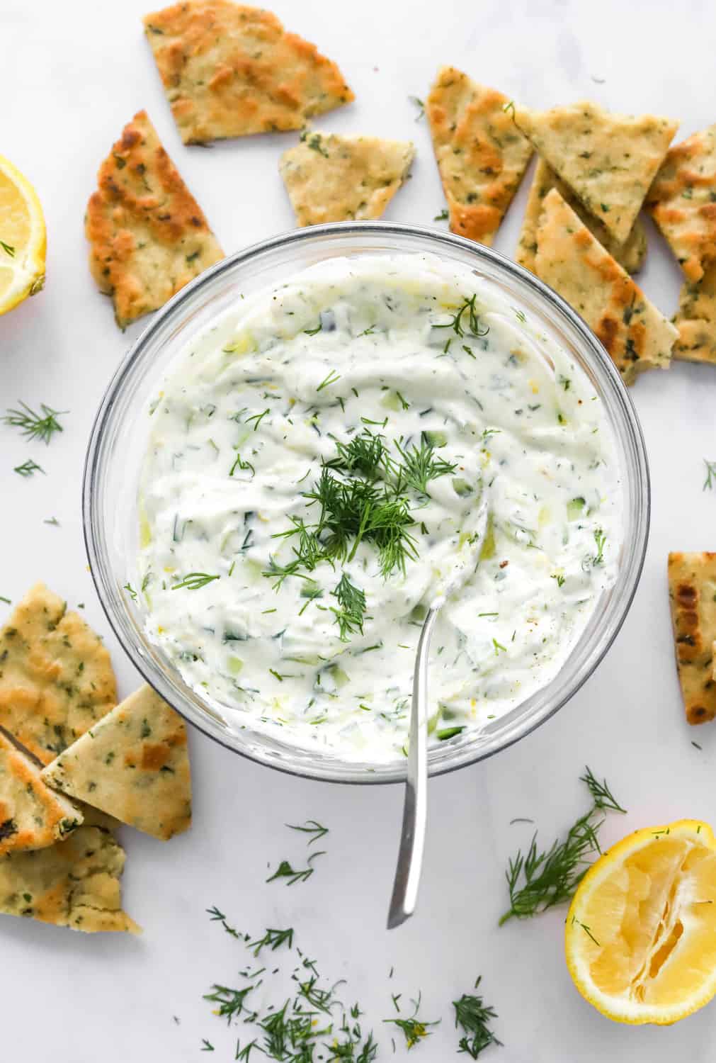 Healthy tzatziki sauce in a round, glass bowl topped with chopped dill with a spoon in the bowl.  With pita chips and lemons around it. 