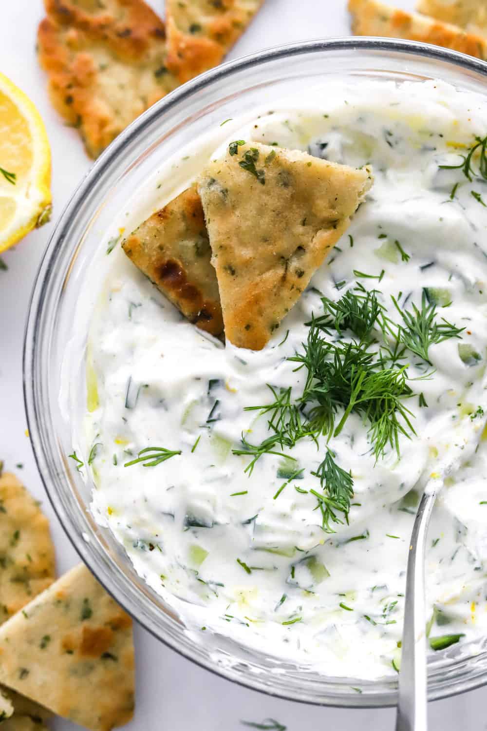 Bowl o Tzatziki sauce with fresh chopped dill on top wit naan wedges in the sauce and more around it. 