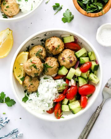 Cooked meatballs in a white bowl with tzatziki sauce and cucumber tomato salad with a slice of lemon next to the bowl and a silver fork on the other side.