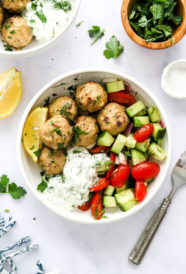 Cooked meatballs in a white bowl with tzatziki sauce and cucumber tomato salad with a slice of lemon next to the bowl and a silver fork on the other side.