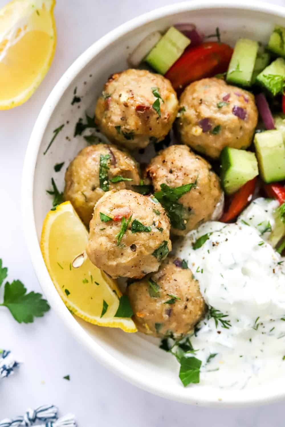 Healthy greek meatballs in a white bowl on top of chopped cucumbers and tomatoes with a creamy white sauce next to them. With a slice of lemon and a few pieces of herbs next to the bowl. 