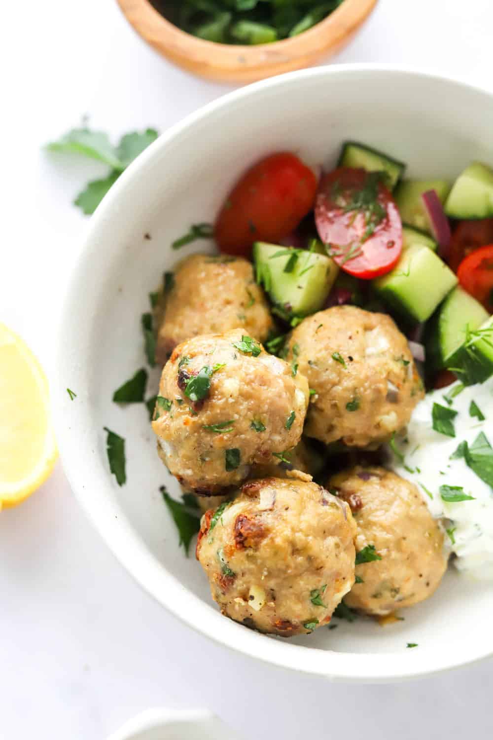 5 golden brown meatballs in a white serving bowl with cucumbers and tomatoes and tzatziki sauce. 