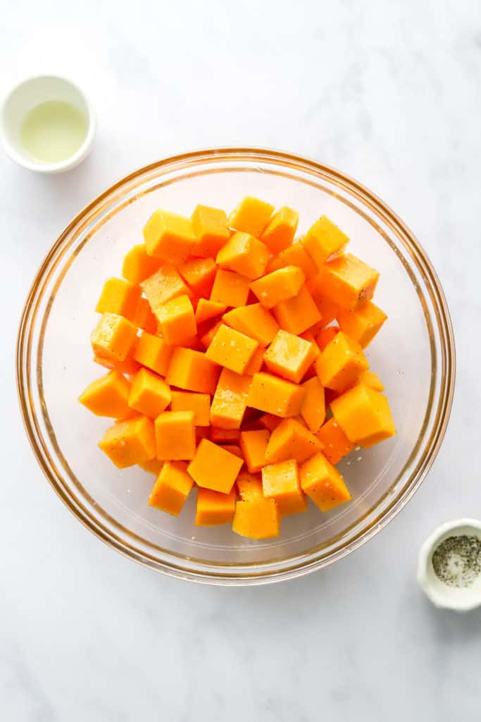 Seasoned, cubed squash in a glass mixing bowl with a smaller bowl of oil behind it and a small bowl of salt and pepper in front of it. 
