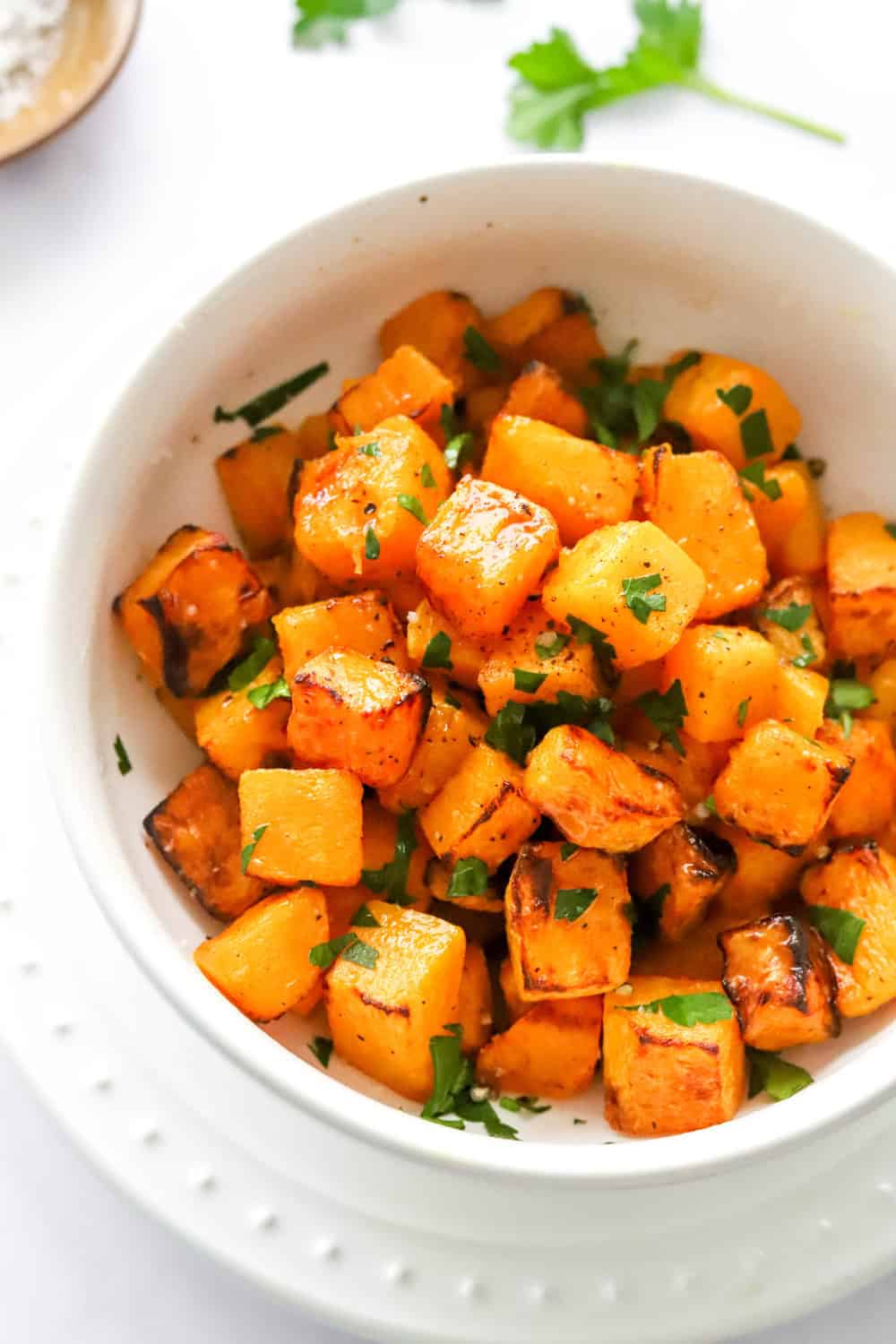 Bowl of cooked cubed squash with chopped green herbs on it in a round, white bowl. 