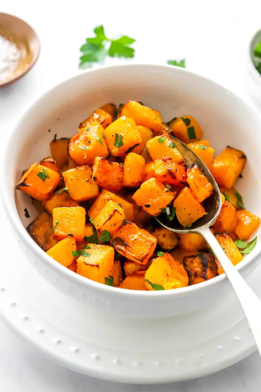 Crispy roasted diced squash in a bowl with a spoon int the bowl and a brown pinch bowl of salt behind it. 