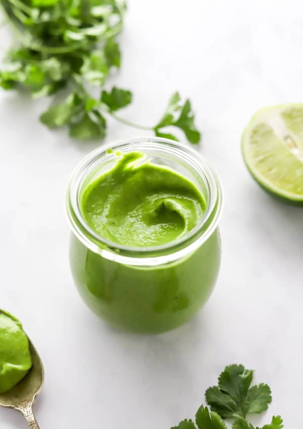 Small glass jar filled with creamy herby cilantro dressing with a cut lime and herbs next to it.