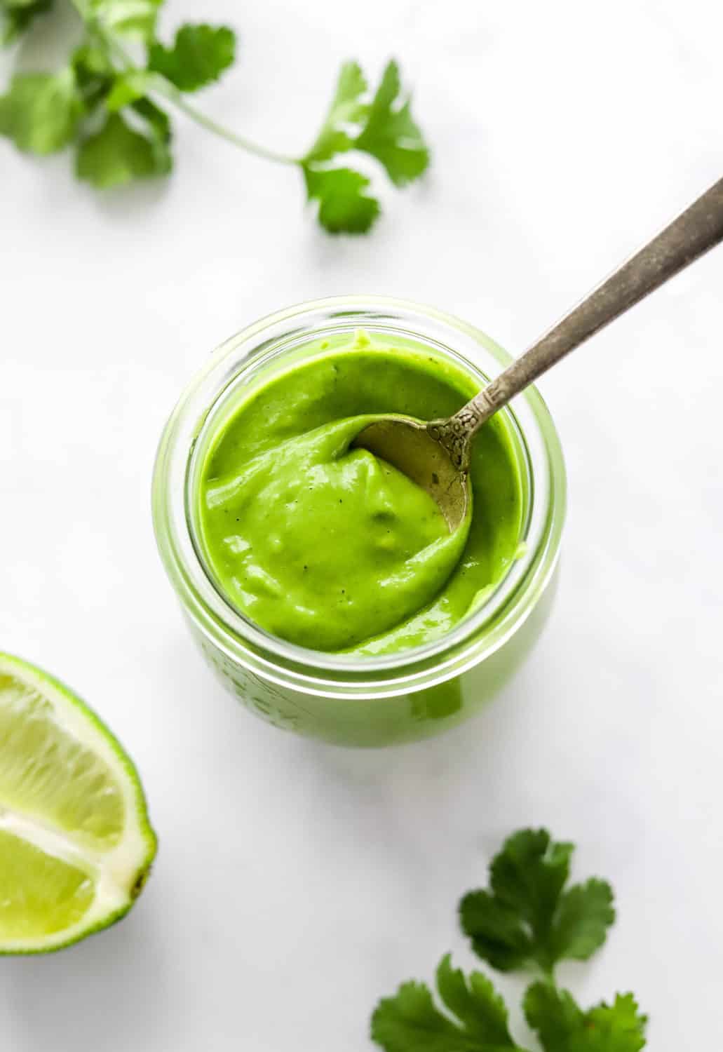 Round, glass jar filled with bright green, creamy dressing with a spoon in the jar.  With a cut lime and torn cilantro in front and in back of it. 