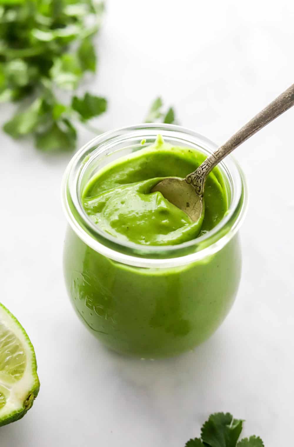 Creamy cilantro lime dressing in a glass jar with a spoon in it with a lime next to it and cilantro leaves behind it. 