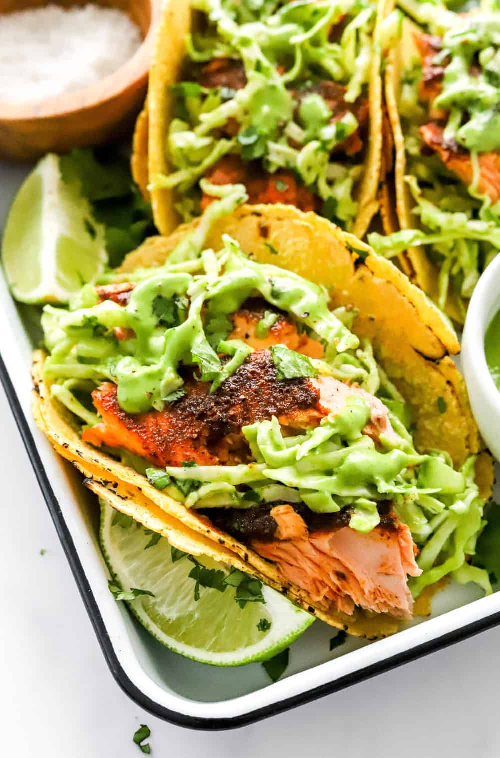 Close up view of cooked, chunks of salmon in a yellow corn tortilla with green dressing and shredded coleslaw on top of the salmon. All on a white and black tray with a lime wedge next to the taco with more salmon tacos behind it. 