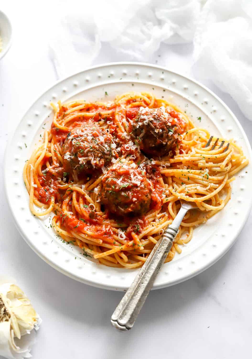 Meatballs layered in red tomato sauce with shredded parmesan on top of them over a bed of cooked spaghetti on a round, white plate. 