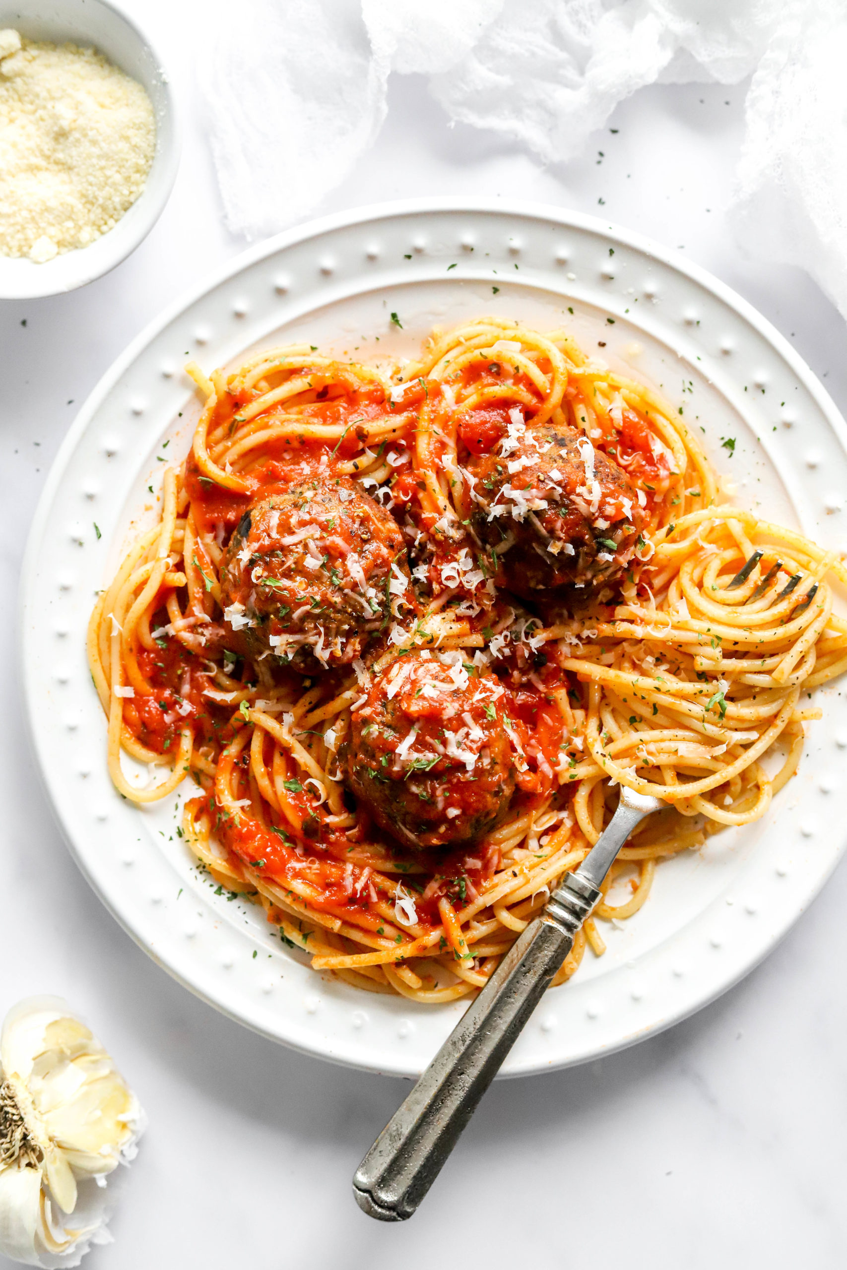 Eggplant meatballs onto of spaghetti covered in tomato sauce on a white plate.  With a bowl of parmesan cheese behind it. 