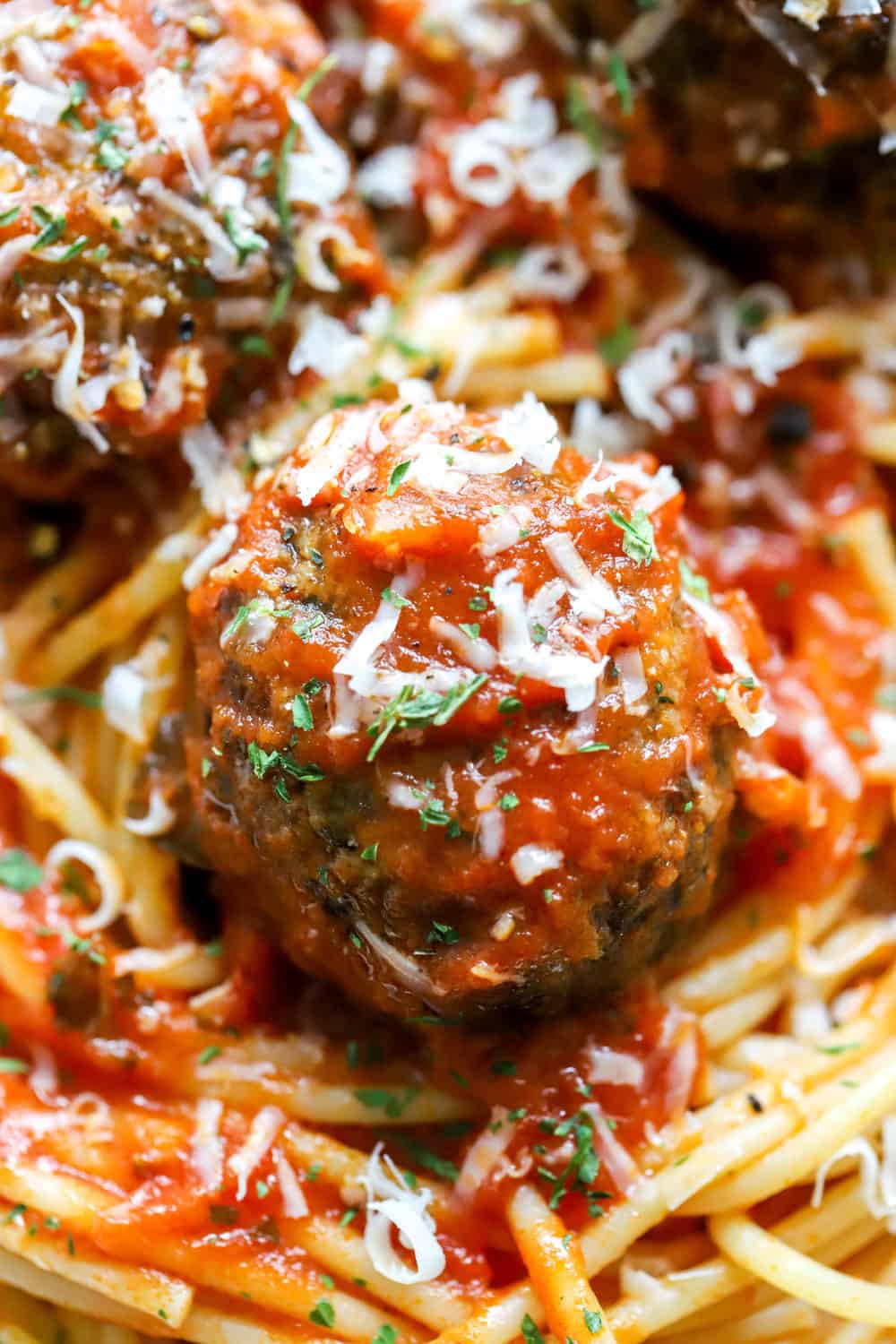 Single meatball covered in spaghetti sauce laying on a bed of cooked spaghetti with cheese and green herbs on it. 