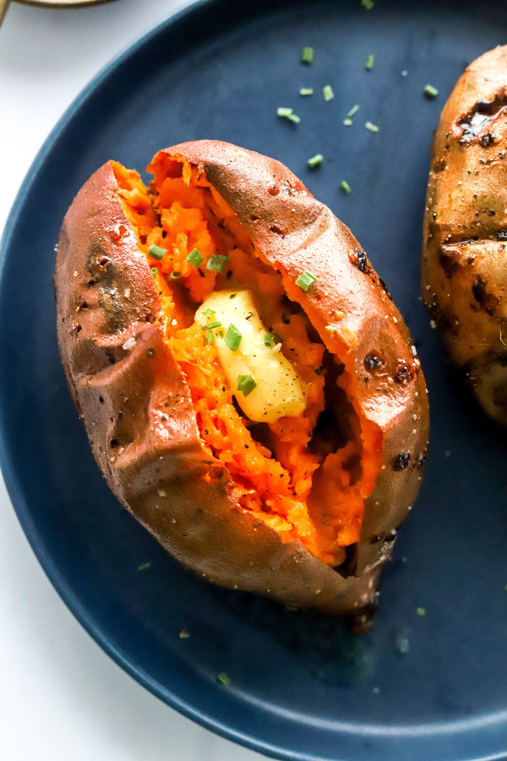 Single sweet potato sliced open with butter in it on a round, blue plate with another baked sweet potato next to it. 