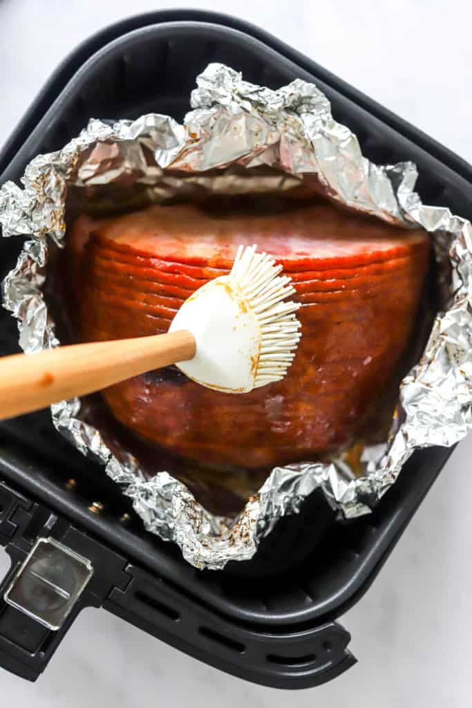 White rubber brush spreading glaze onto a cooked ham in some foil in an air fryer basket. 