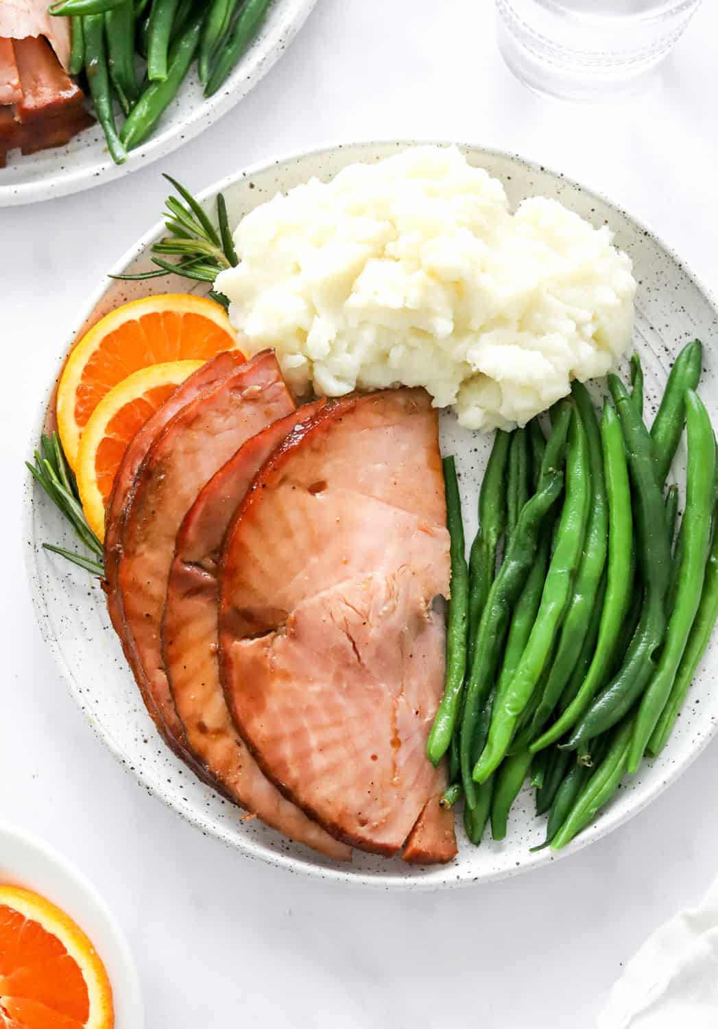 Sliced, cooked air fryer ham on a round plate with green beans and mashed potatoes with orange slices on the plater and in front of it. 