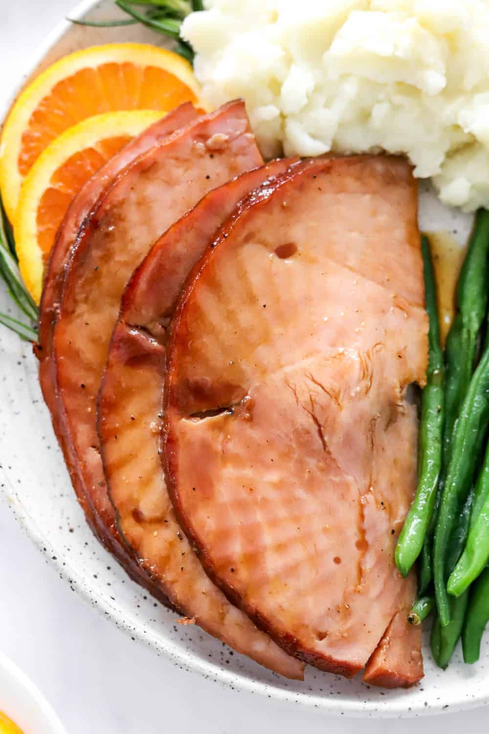 4 slices of glazed ham on plate with green beans and mashed potatoes around it with a few orange slices under it. 