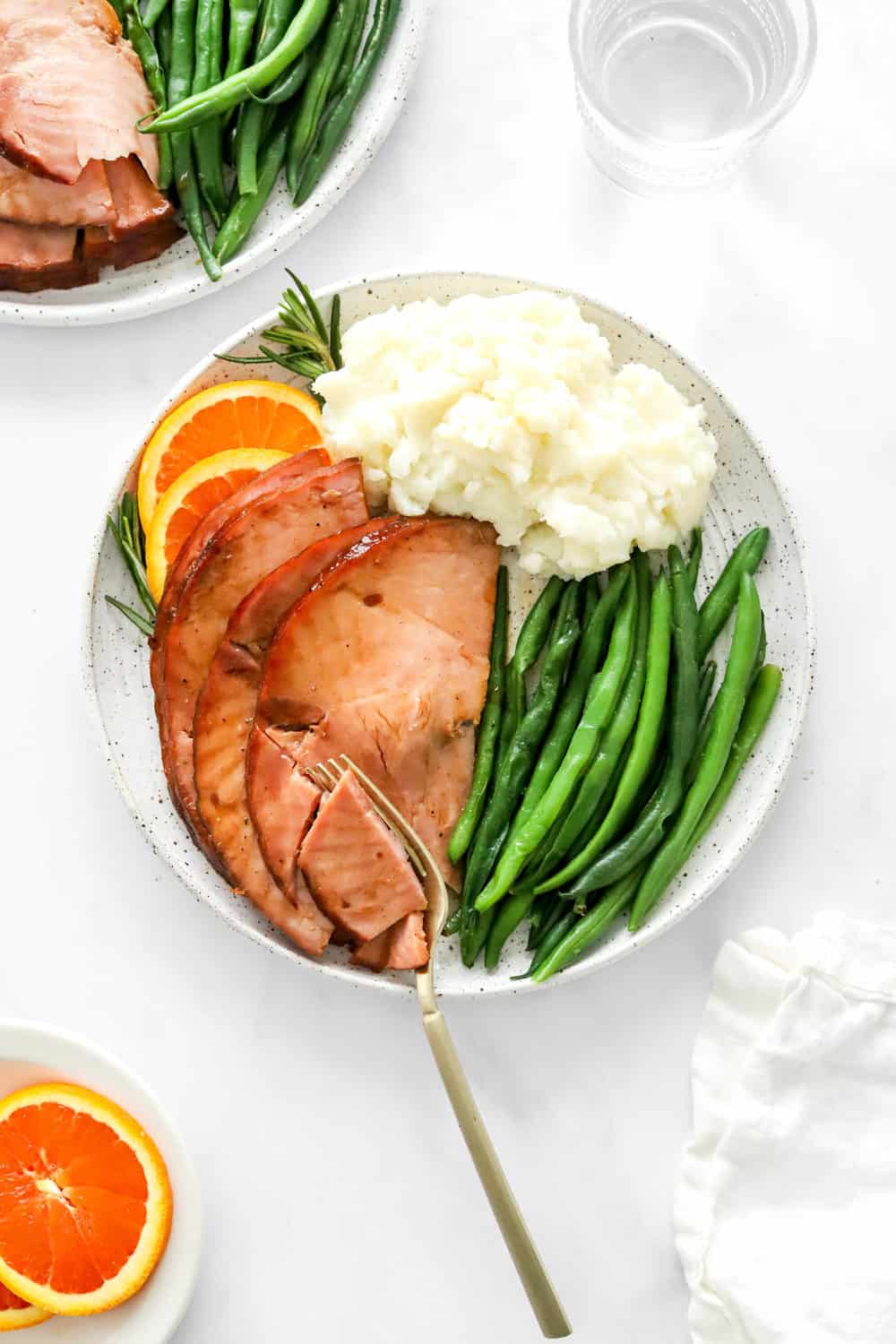Ham slices on a round plate next to mashed white potatoes and string green beans with a fork on the plate. 