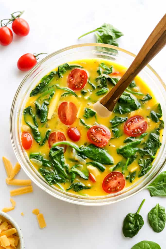 Whisk eggs in a bowl with spinach and tomatoes with a wooden spatula in the bowl. 