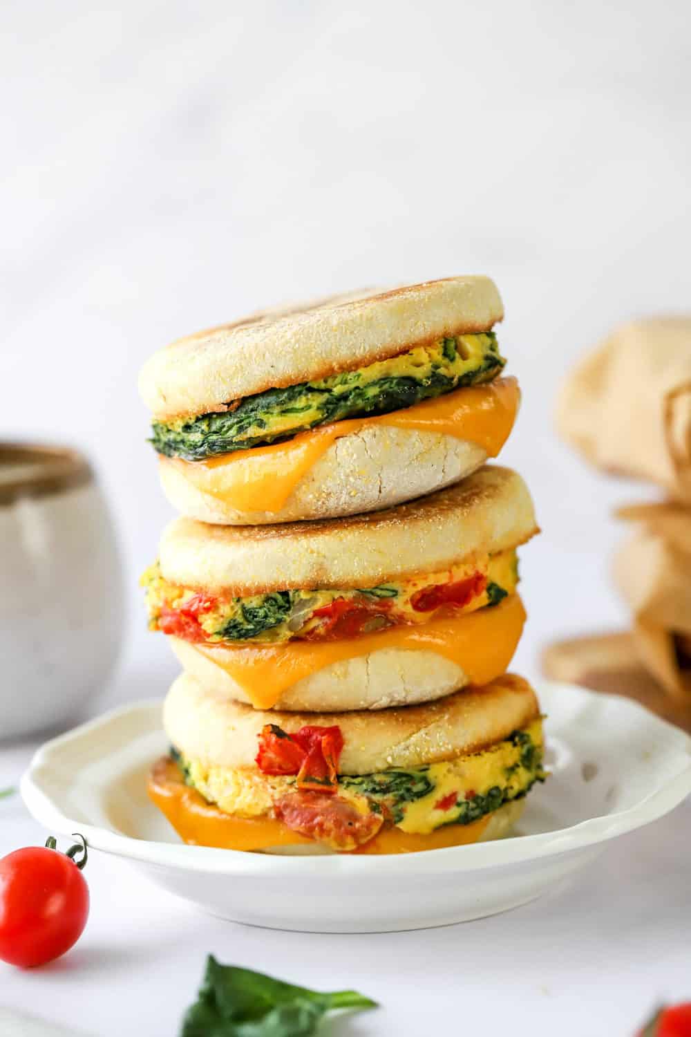 Three breakfast sandwiches stacked on top of one another on.a plate with wrapped sandwiches and a cup of coffee behind it. 