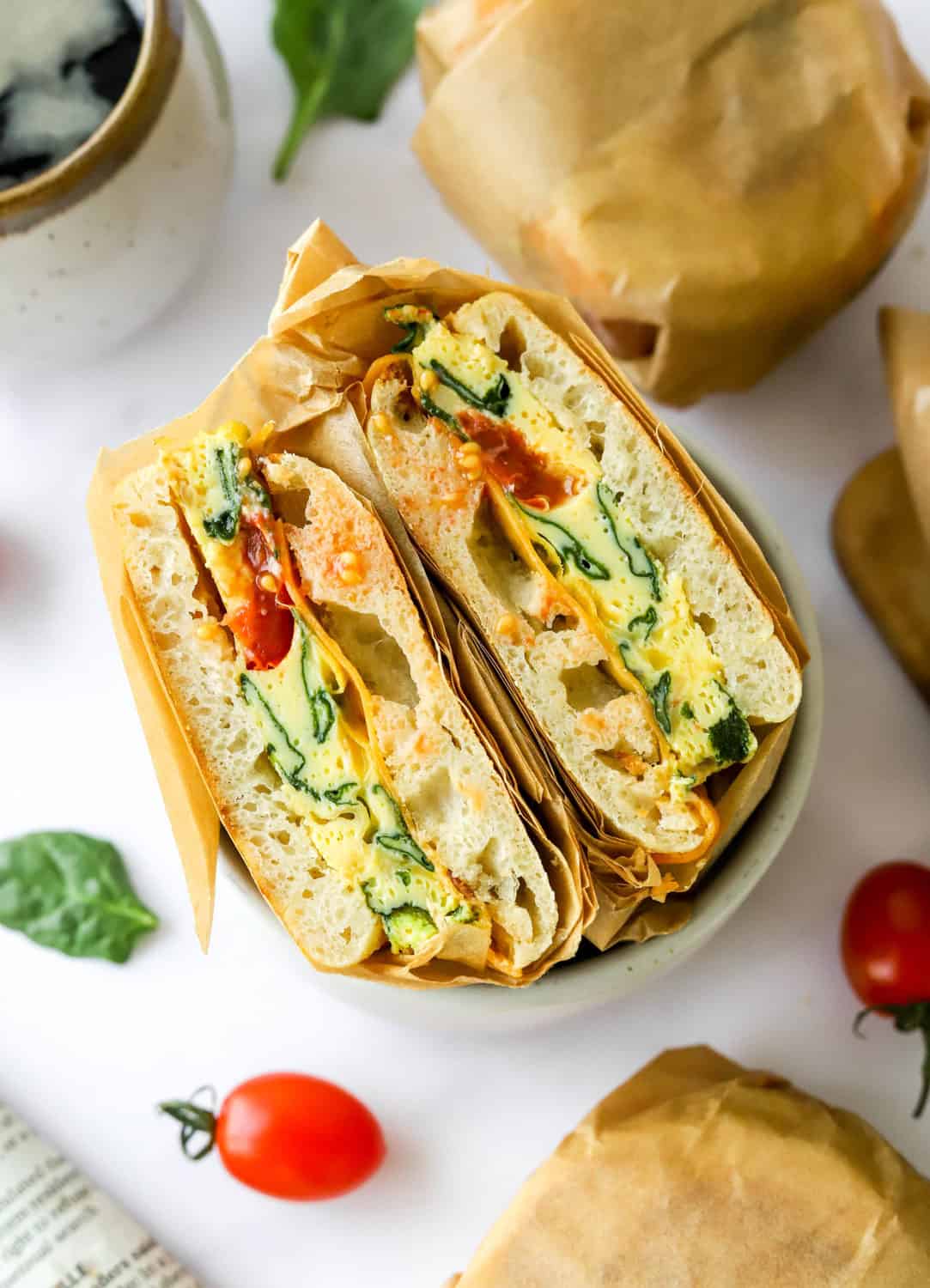 Make ahead breakfast sandwiches wrapped in brown parchment paper with one sliced open on a plate and more wrapped sandwiches behind it. 