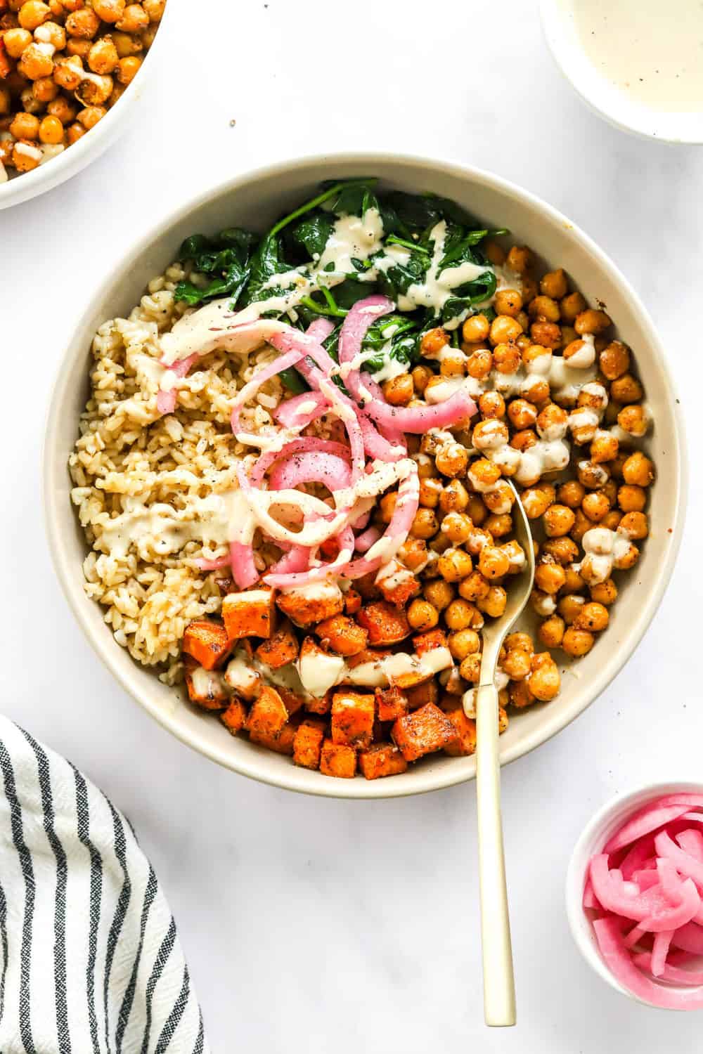 Round bowl filled with sweet potatoes, chickpeas, brown rice and greens.  Topped with a white dressing with a spoon in the bowl. 