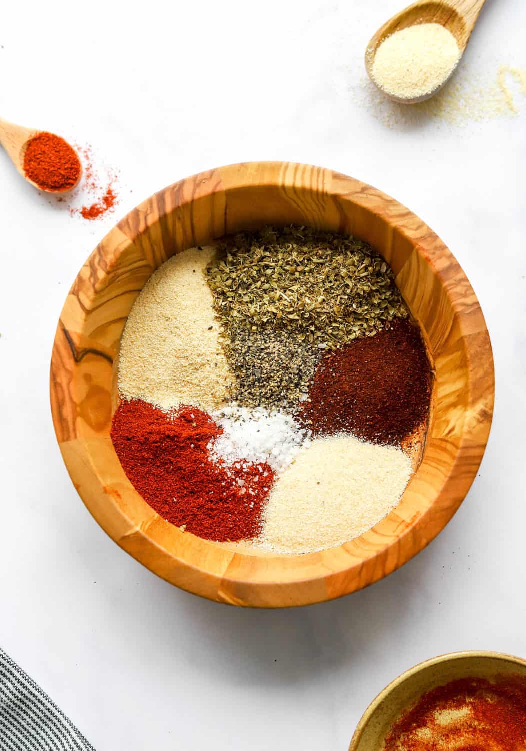 Spices separated in a round, wooden bowl with a small wooden spoon with paprika in it behind it. 