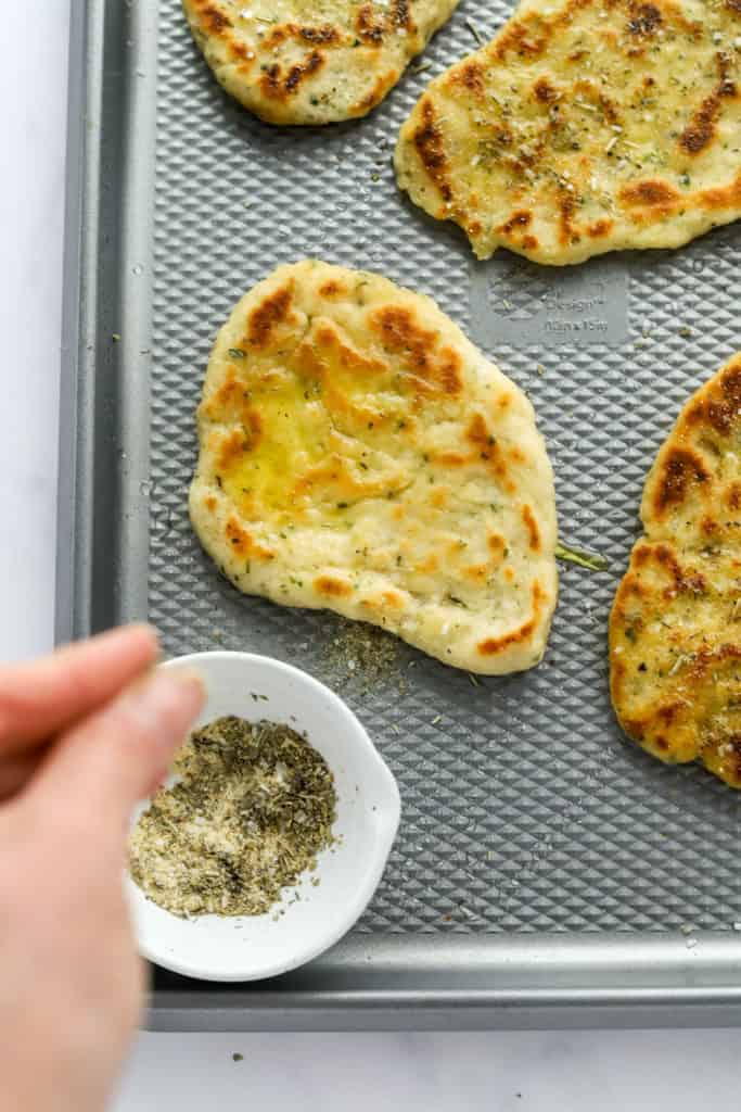 Golden naan bread on a baking sheet with a hand sparkling seasoning on it. 