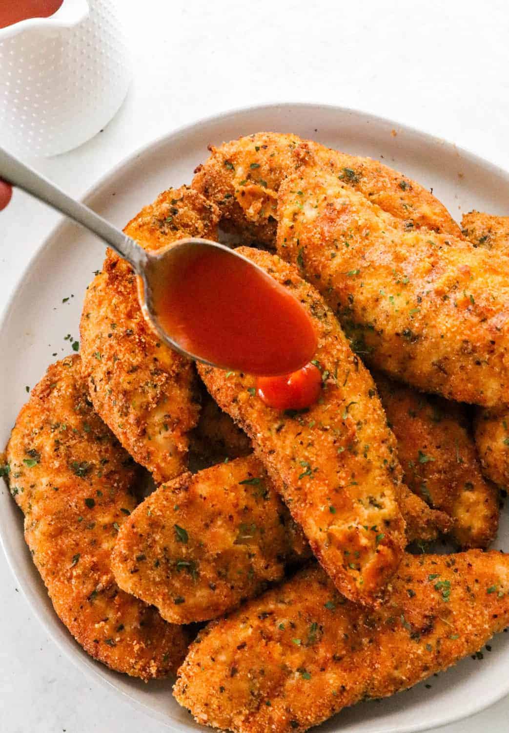 Spoon dripping buffalo sauce onto crispy breaded chicken tenders. With more hot sauce in a white jar behind it. 