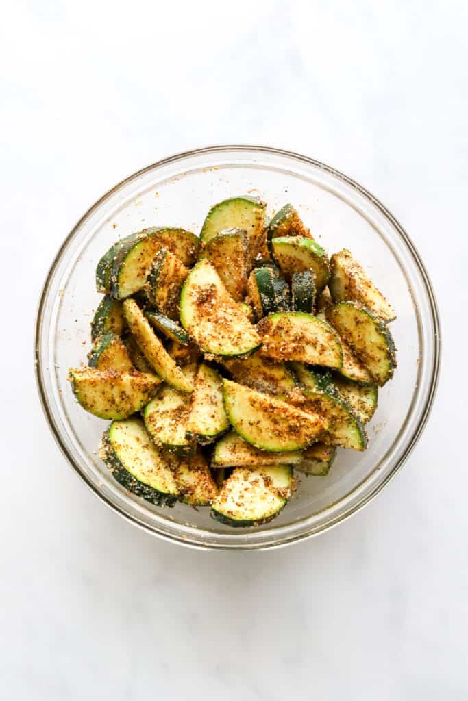 Sliced Zucchini in a glass mixing bowl with seasoning on it. 