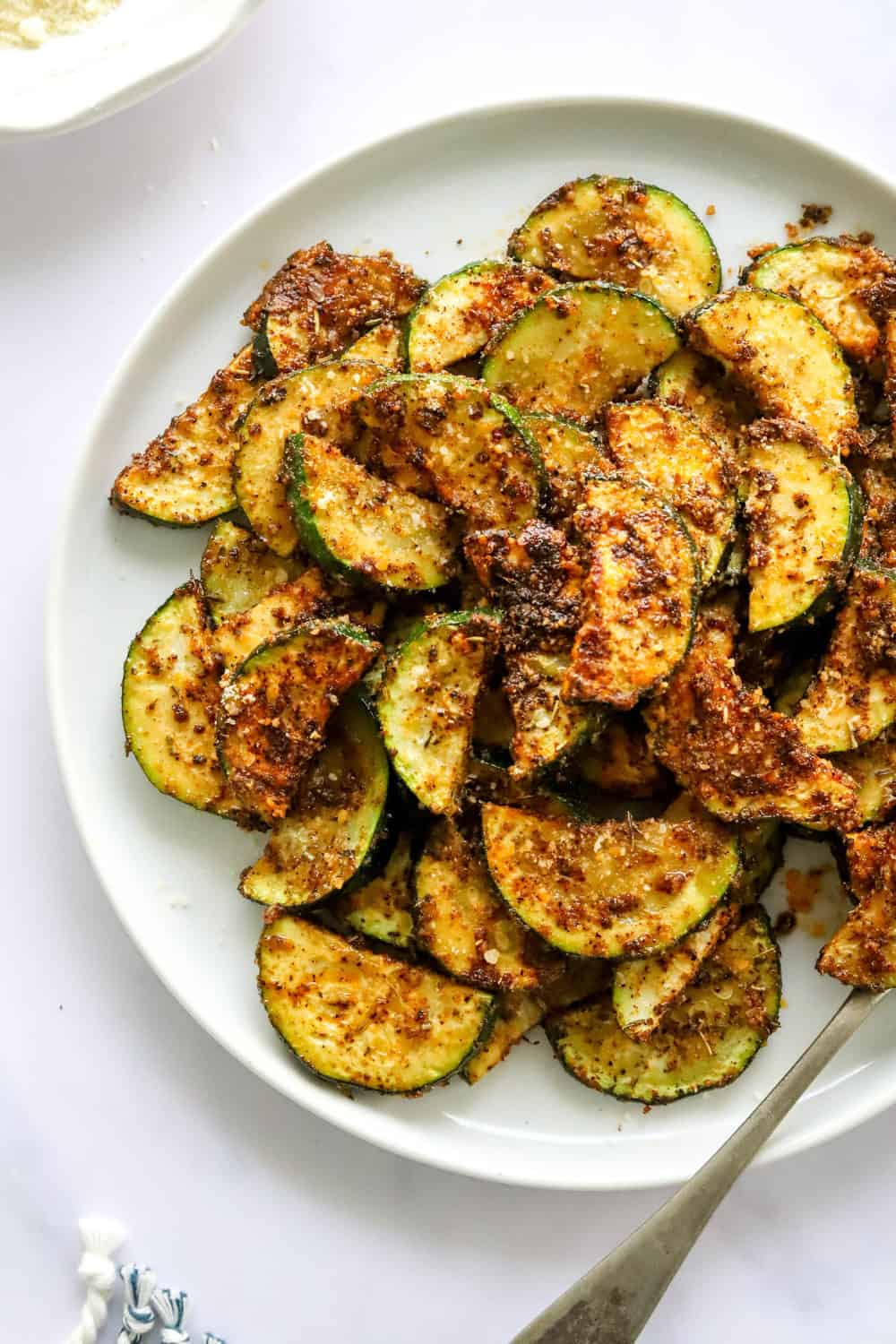 Sliced, seasoned and roasted crispy zucchini on a white plate with silver spoon on the plate. 