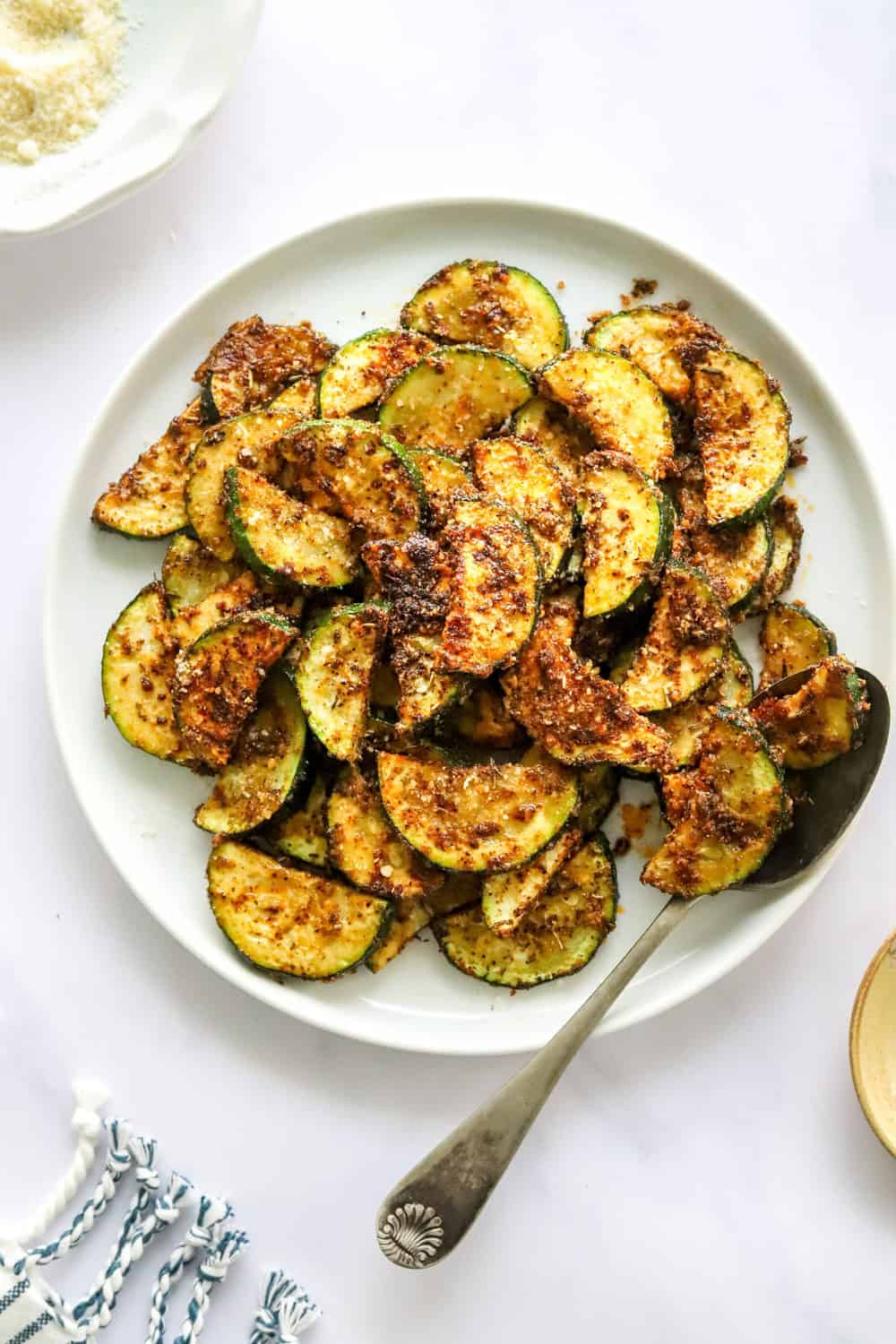 Round, white dinner plater filled with sliced, air fried zucchini with a spoon on the plate with zucchini on it. 