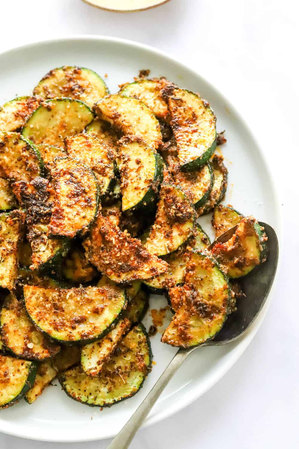 Piled of roasted zucchini with seasoning on it on a white plate with a spoon on the plate. 