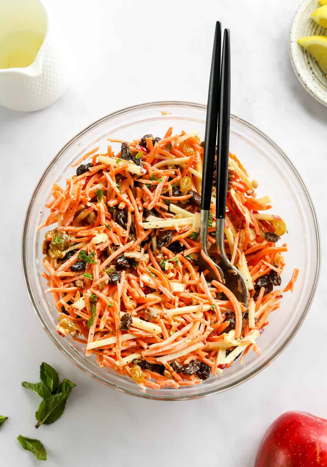 Carrot raisin salad in a round glass mixing bowl with black and silver serving spoons in the bowl. 