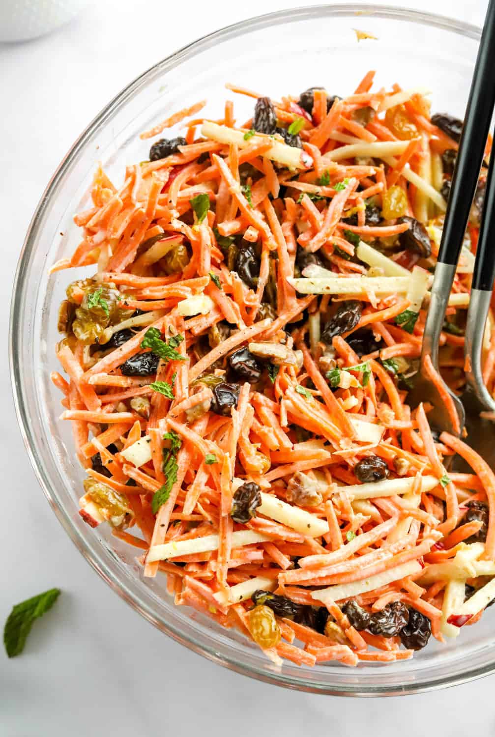 Shredded carrot salad in a glass bowl with raisins, nuts and mint with some shredded apple as well.  With black serving spoons in the bowl. 