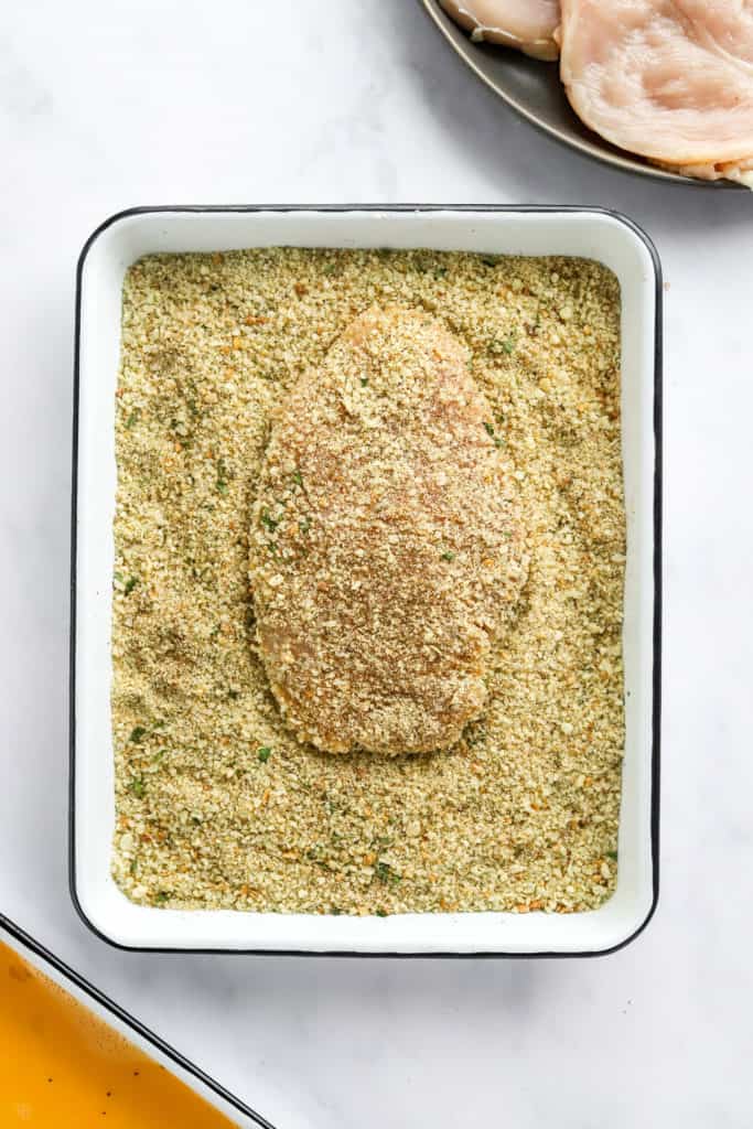 Uncooked, breaded chicken breast in a shallow white dish of breadcrumbs. 