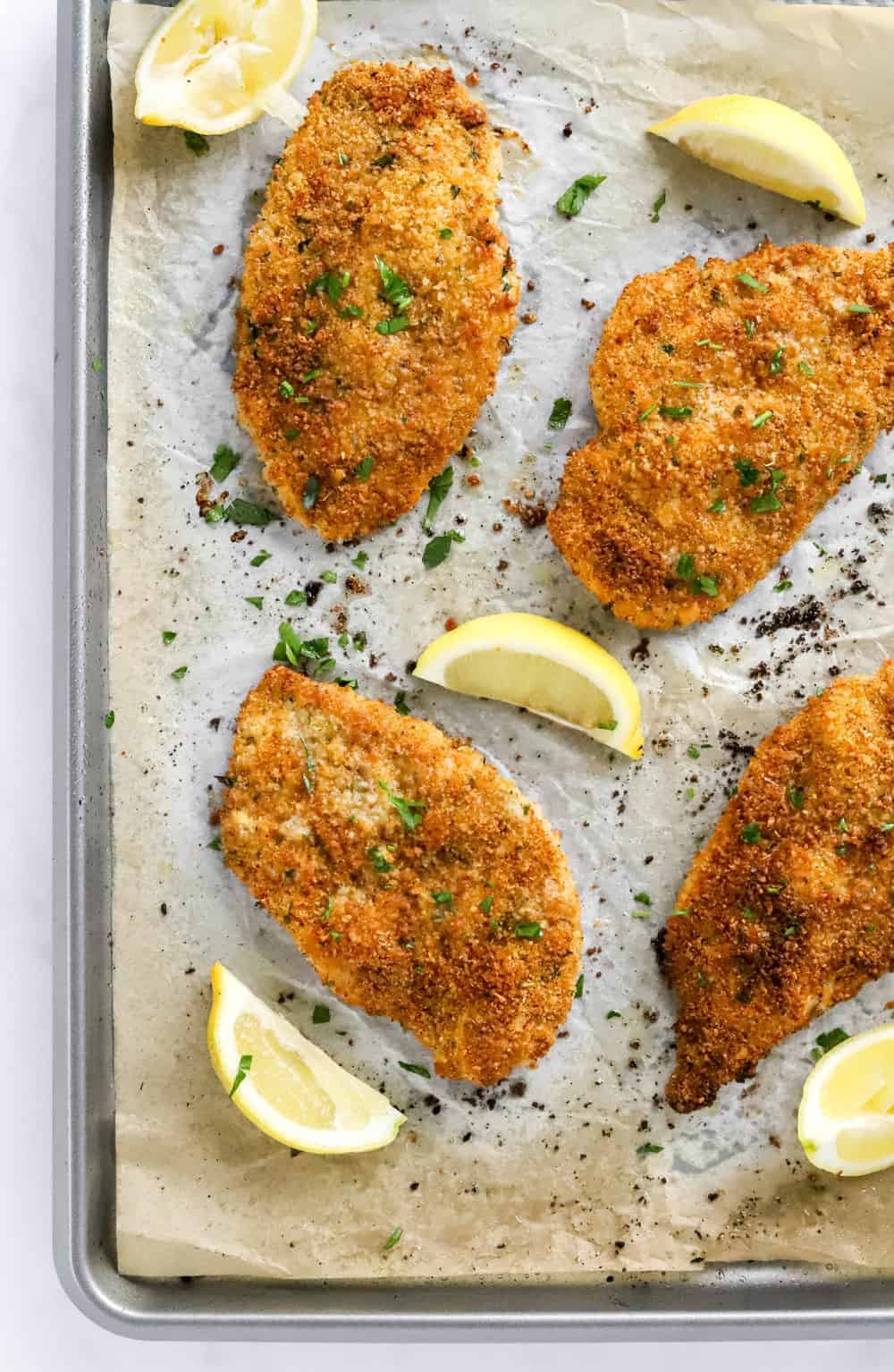 4 pieces of oven fried chicken on a baking sheet with herbs sprinkled on them and lemon wedges around them. 