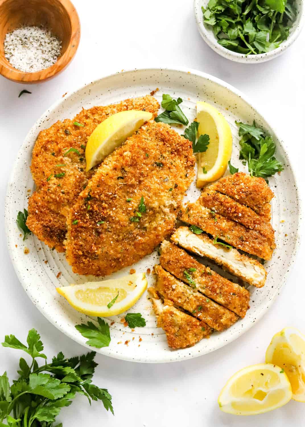 Round white specked plate with breaded chicken cutlets on it with one sliced with lemon wedges and parsley leaves around it. 