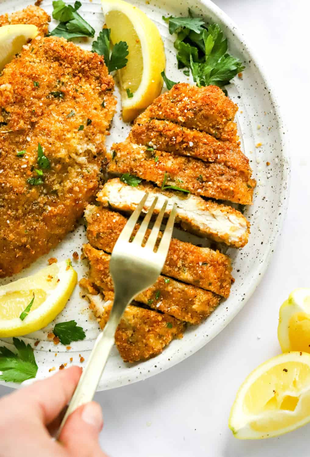 Sliced breaded chicken on a plate with more chicken next to it and a hand picking up a piece of chicken with a gold fork. 