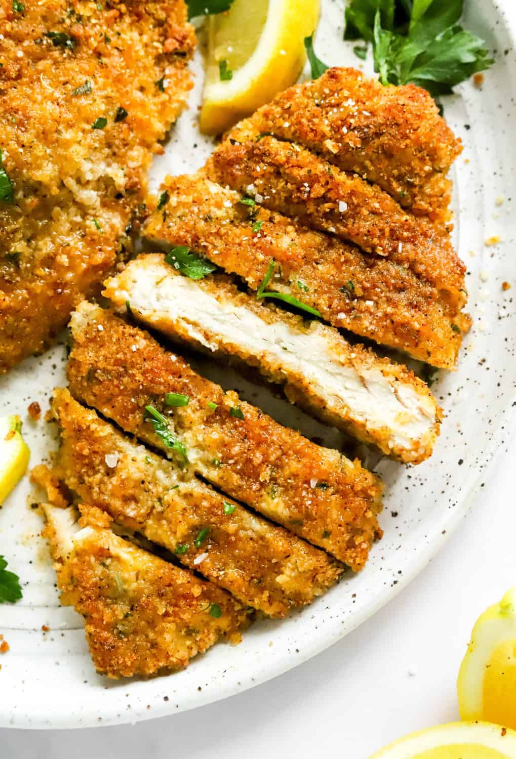 Crispy breaded, baked chicken sliced into long pieces on a white plate with more chicken next to it and a lemon slice in front of it. 